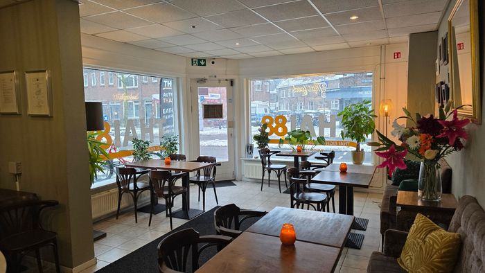 Interieurfoto van een gezellig, warm verlicht restaurant met meerdere eettafels, houten stoelen en grote ramen aan de straatkant.