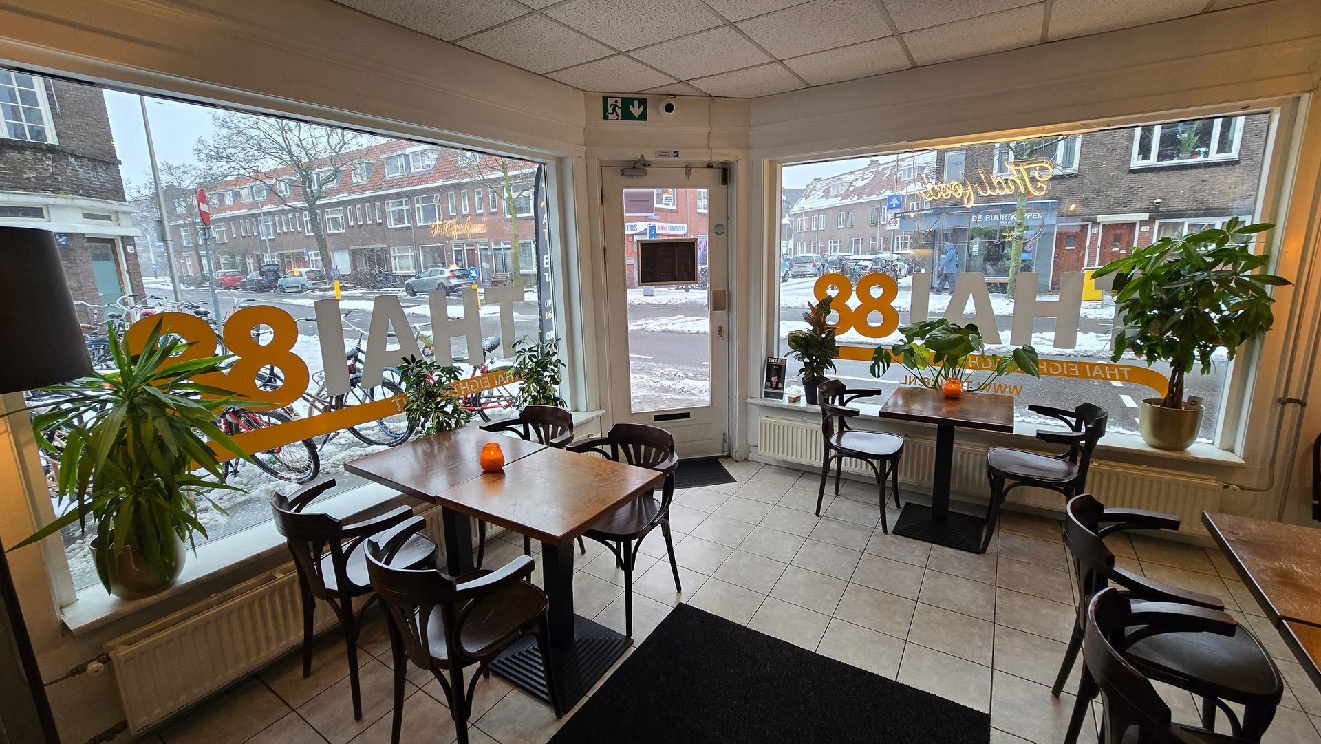 Binnen in een helder verlicht café met grote ramen die uitkijken op een besneeuwde straat, met houten tafels, stoelen en potplanten.
