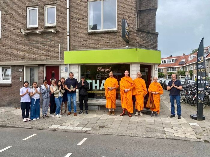 Een groep mensen, waaronder drie boeddhistische monniken in oranje gewaden, staat voor een winkelpand met een groen uithangbord.
