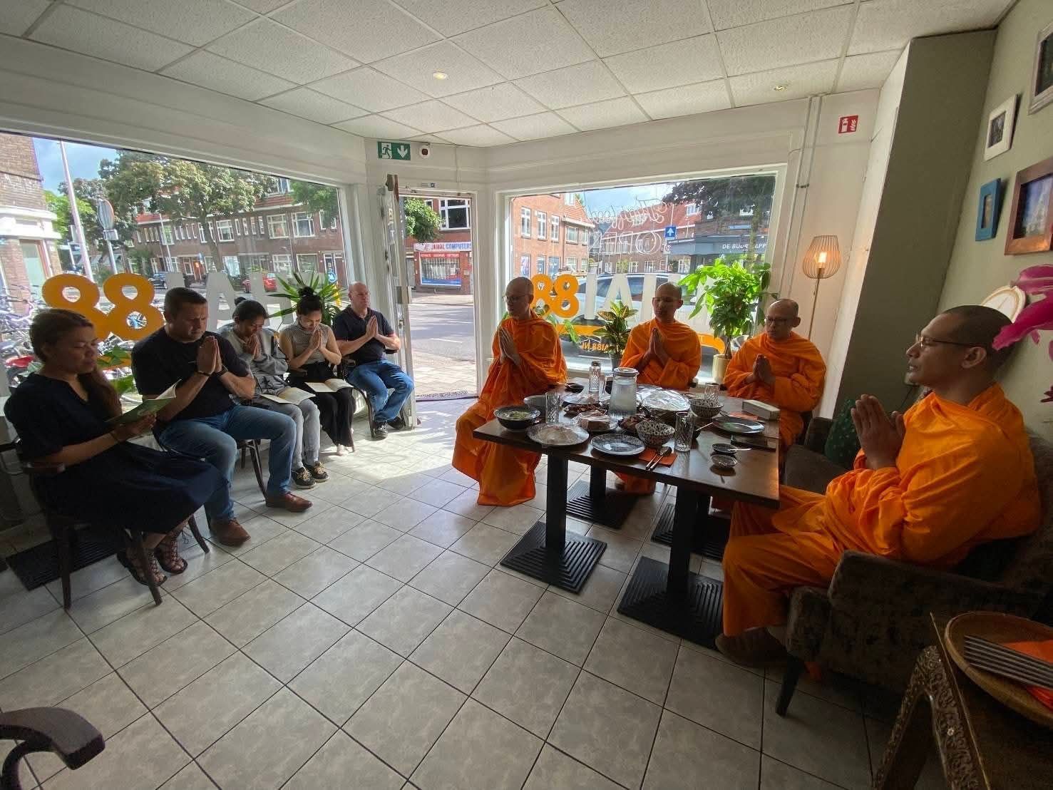 Een groep mensen en monniken in oranje gewaden zitten samen aan een tafel in een lichte ruimte, met de handen gevouwen in gebed of meditatie.