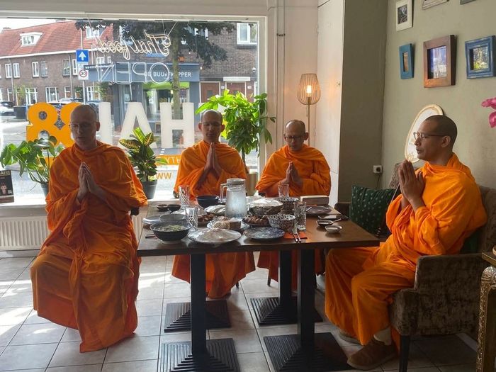 Vier monniken in oranje gewaden zitten aan een tafel in een felverlichte ruimte, met hun handen in een gebedshouding.