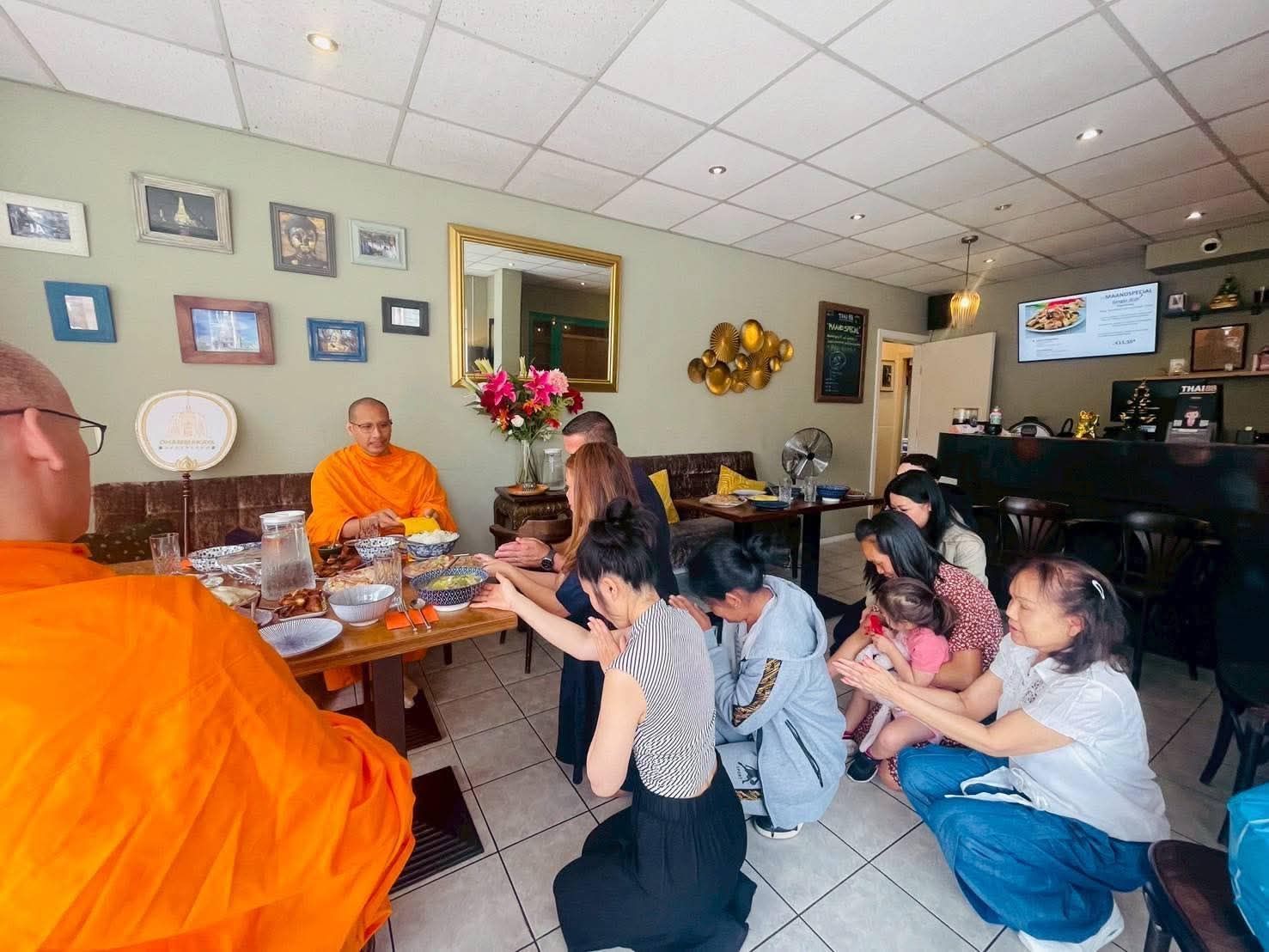 Mensen in vrijetijdskleding knielen op de grond voor een monnik in oranje gewaden en nemen deel aan een religieuze ceremonie.