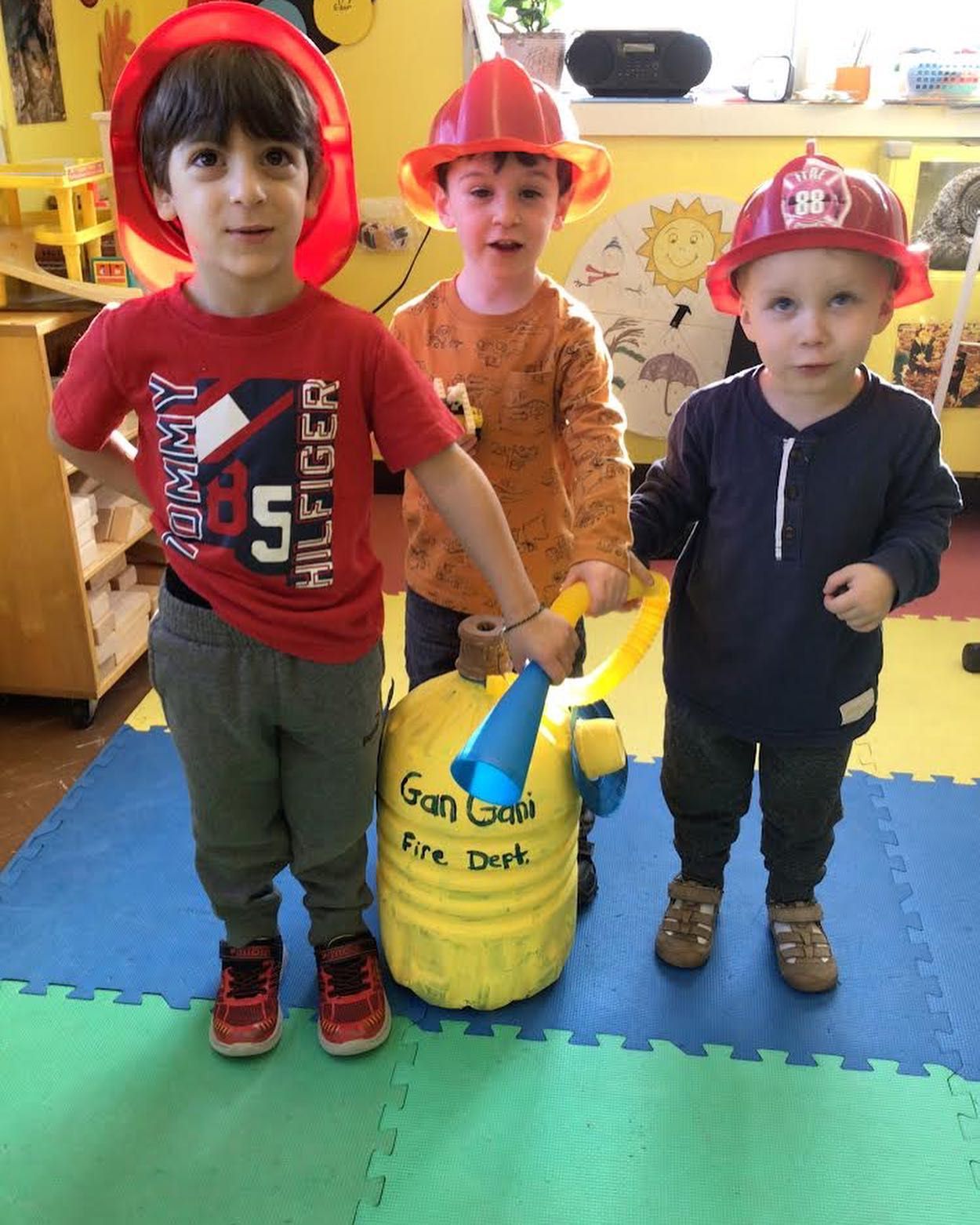 Three young boys wearing fireman hats and holding a toy fire hydrant