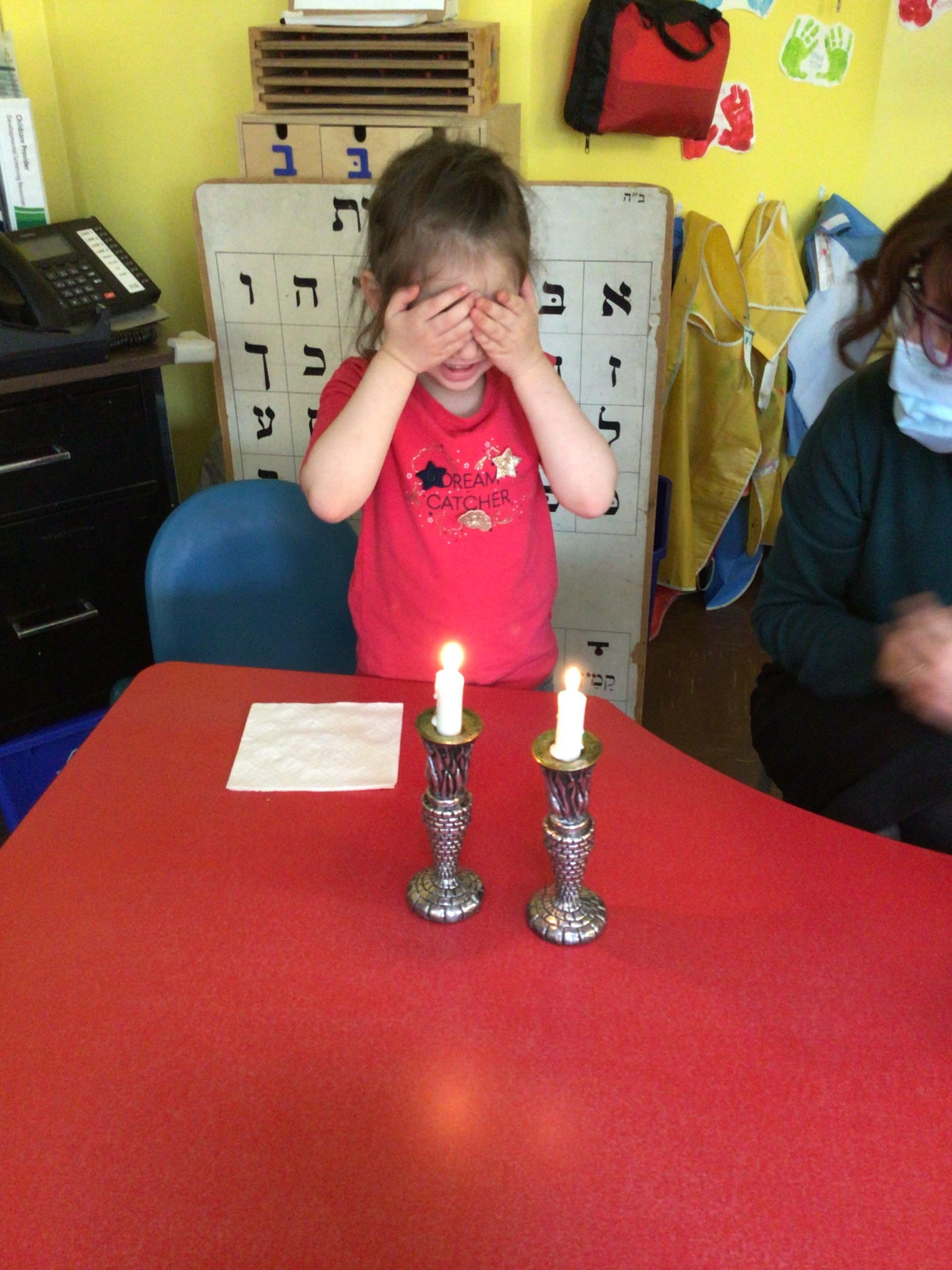 A little girl covering her eyes while sitting at a table with two candles