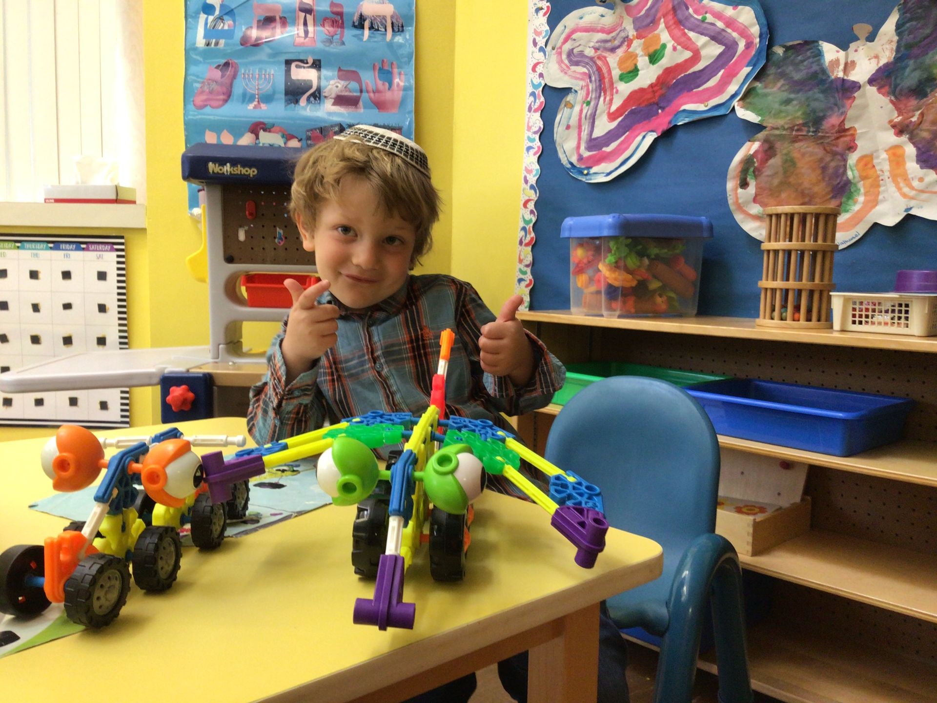 A young boy is sitting at a table with toys and giving a thumbs up