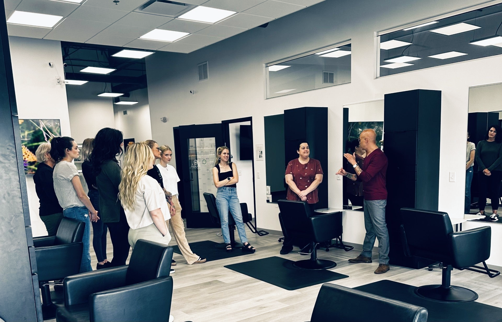Group of people in a salon, listening to a person speaking, standing near chairs and mirrors.