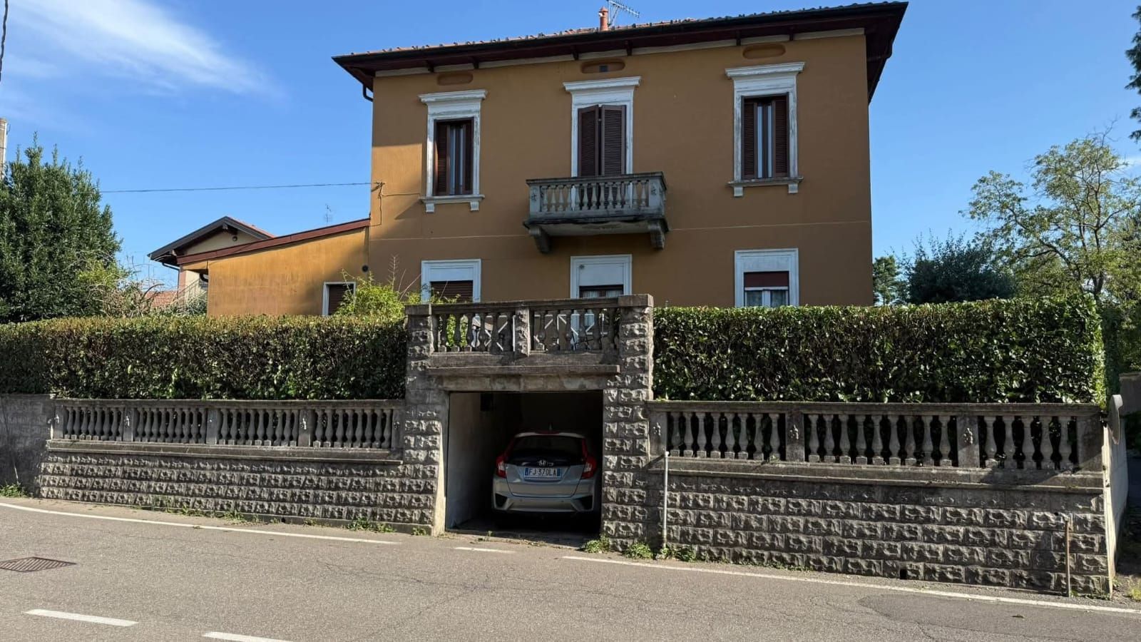 Casa gialla a due piani con balcone, siepe e muro in pietra; un'auto è parcheggiata all'ingresso del garage.