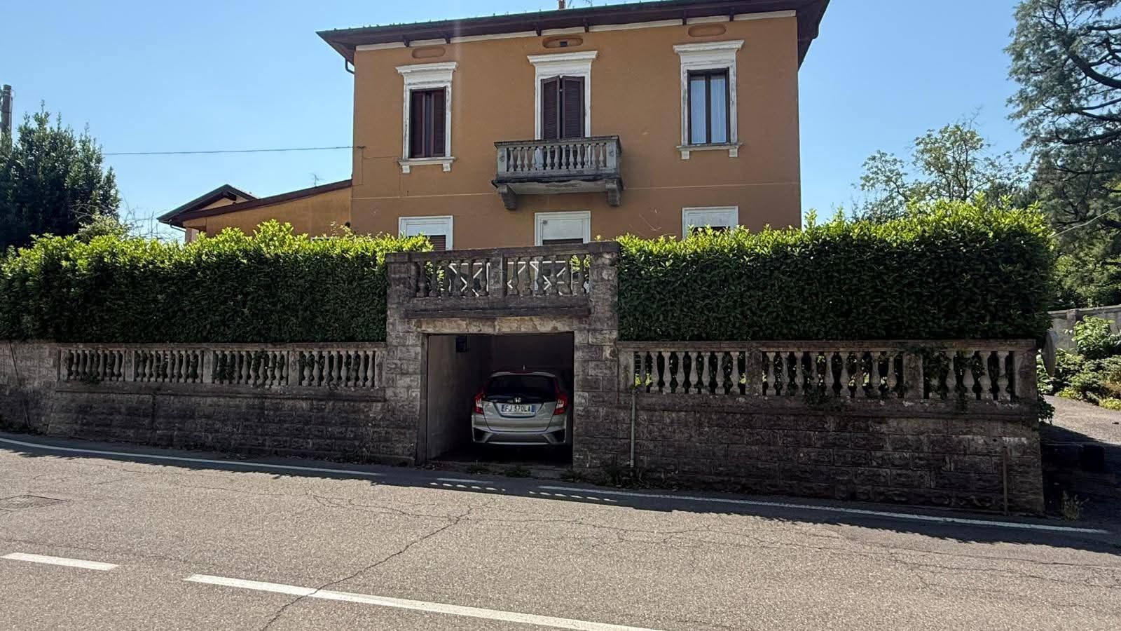 Casa color sabbia a due piani con balcone e un'auto parcheggiata in un posto auto coperto dietro un muro di mattoni e una siepe verde.