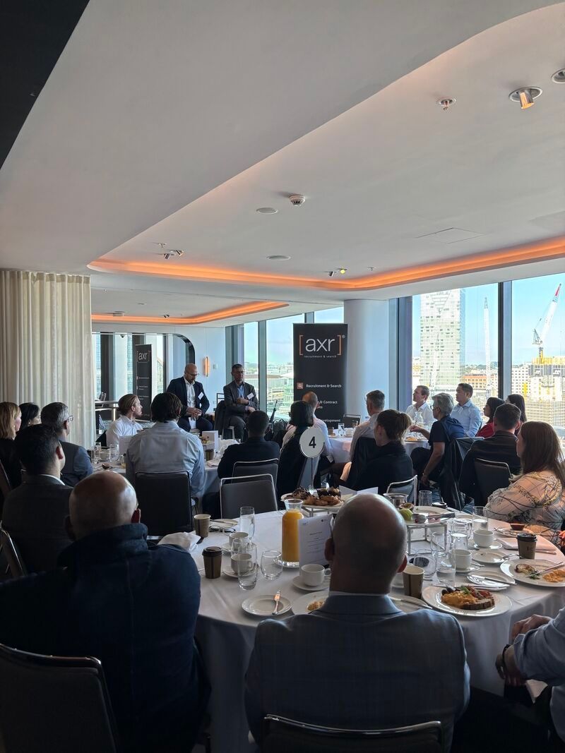 People at a conference listening to a panel discussion in a light-filled room with round tables.