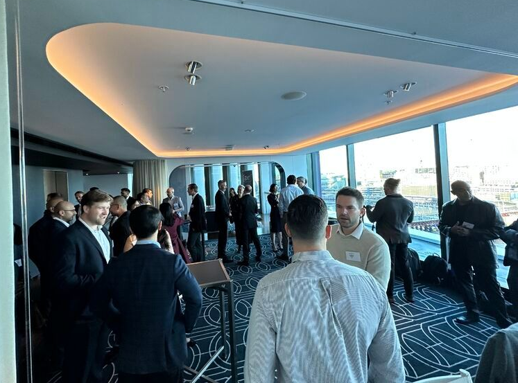 People networking in a well-lit room with large windows overlooking a city, some are conversing.