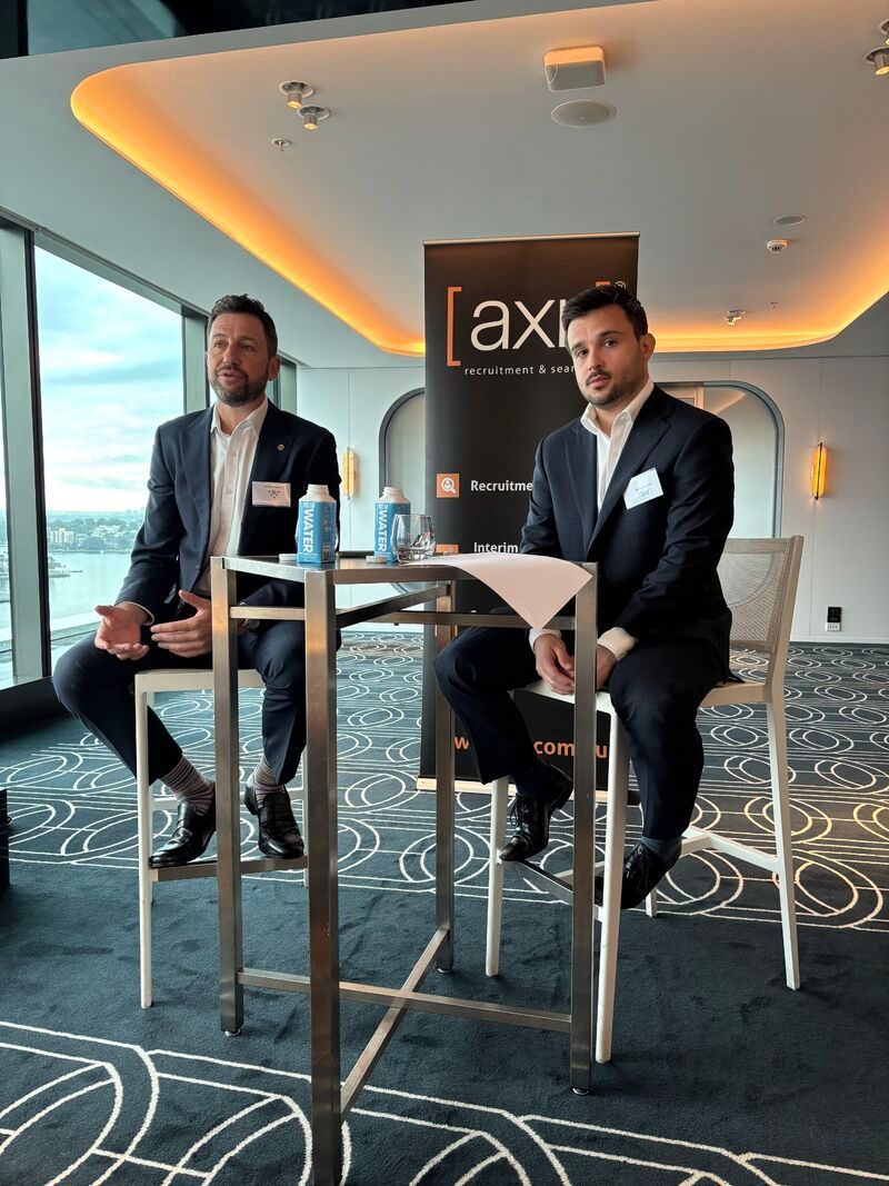Two men in suits seated at a table on high chairs, presenting at an event, with a cityscape background.
