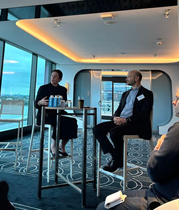 Elon Musk speaking at a panel, seated with two others at a high-top table, in a well-lit room with city view.