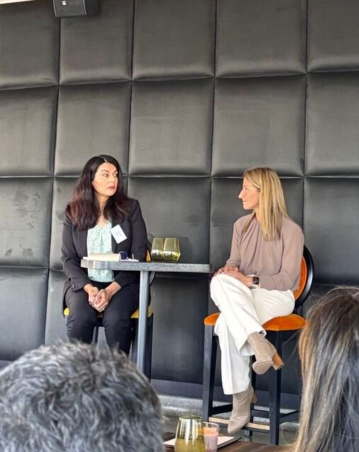 Two women seated at a table, panel discussion. One speaks, another listens. Dark background, audience in foreground.