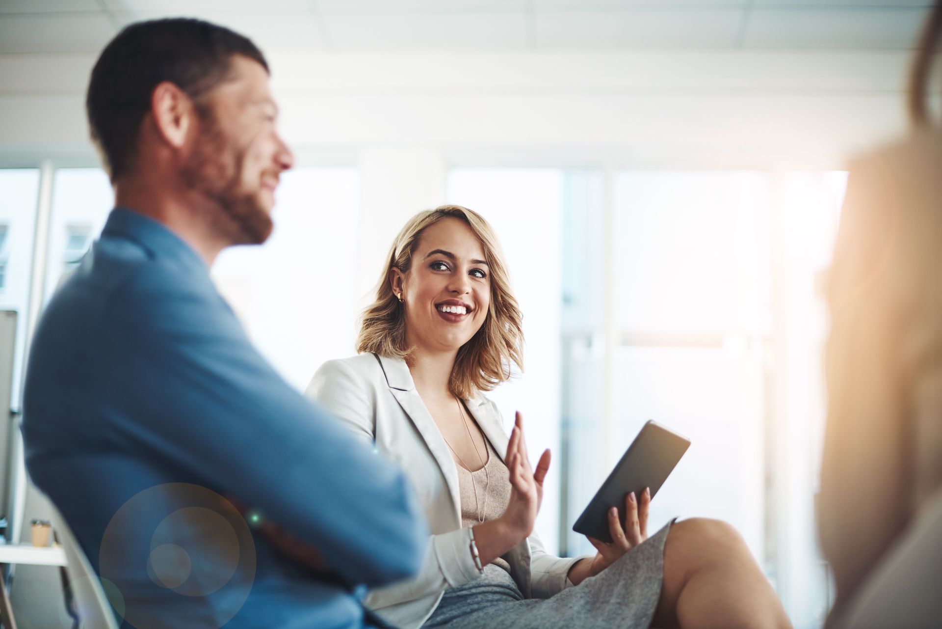 Woman holding tablet gestures while smiling at a man with crossed arms in an office setting.