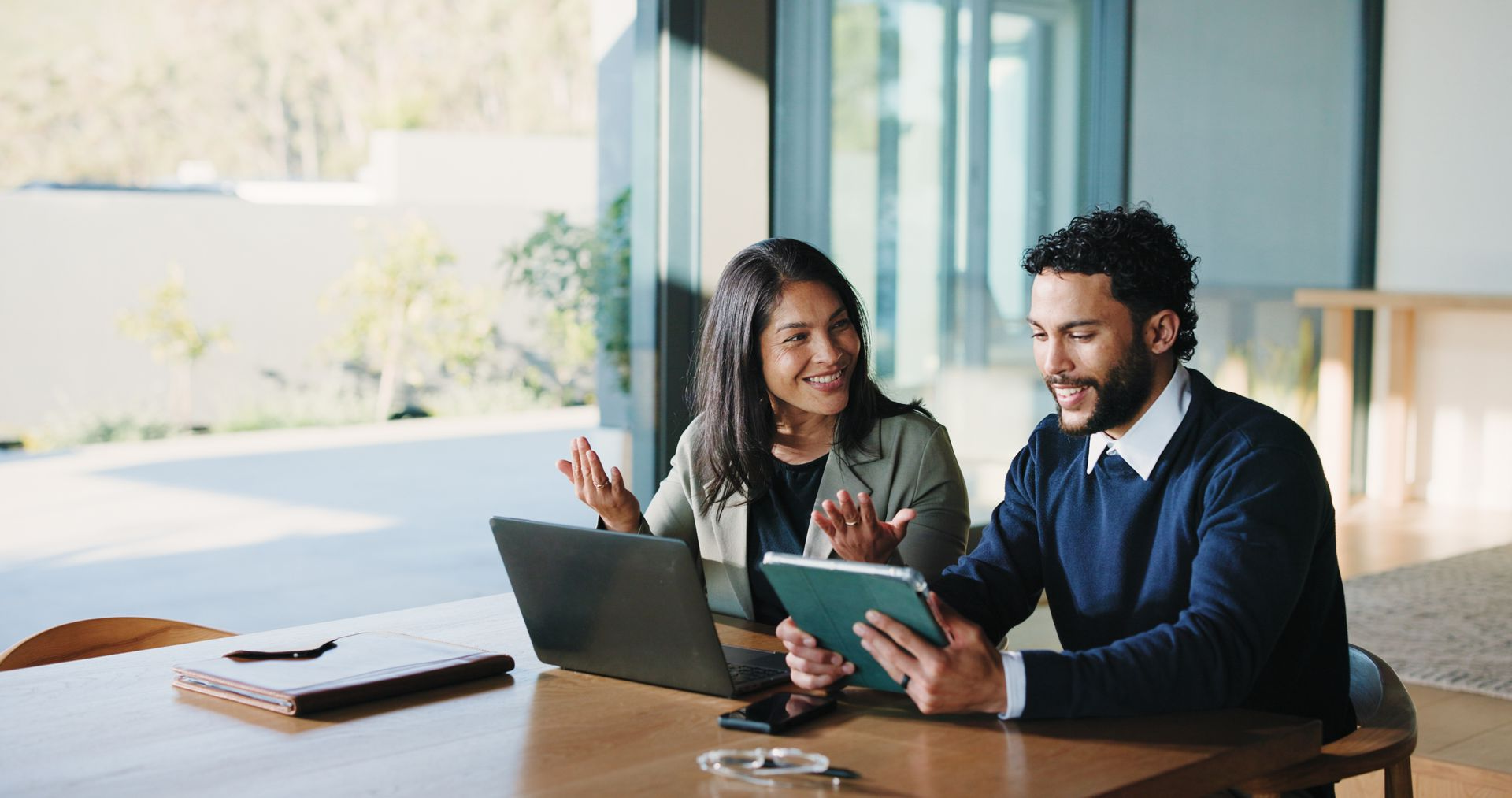 Two people at a table, using a laptop and tablet, smiling and collaborating in a bright room.