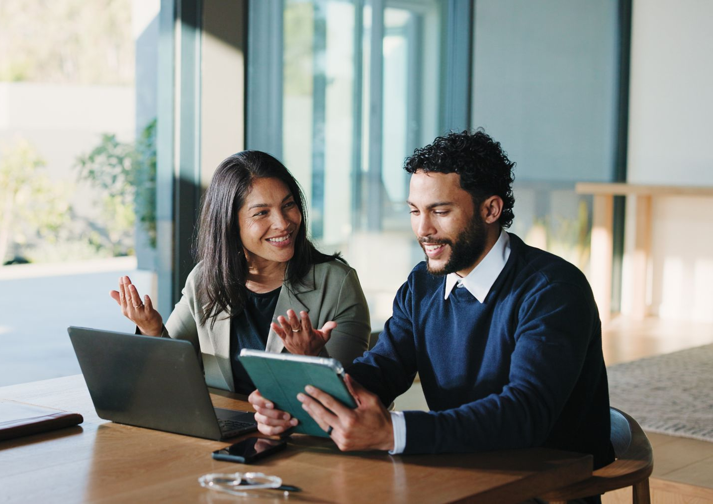 Two people at a table, using a laptop and tablet, smiling and collaborating in a bright room.
