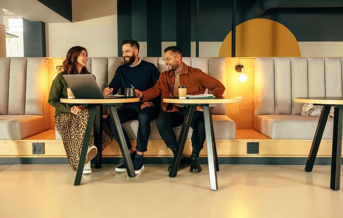 Three people in casual attire seated around a small table in a modern, neutral-toned office space, looking at a laptop.