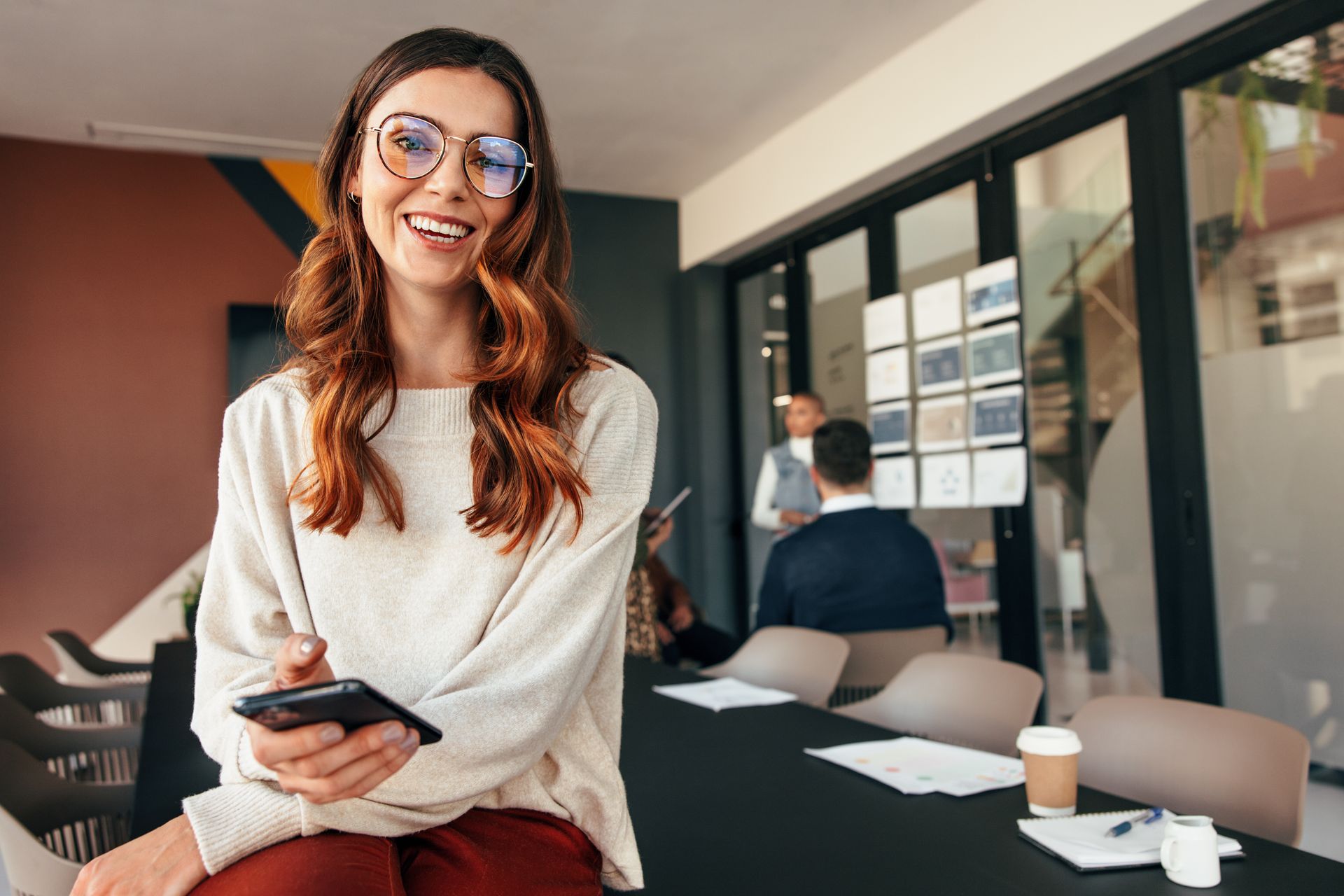 Woman in glasses smiles, holding phone, seated on table. Meeting in the background.