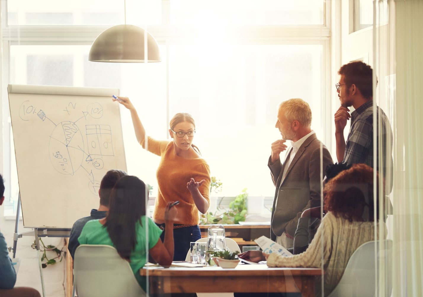 Woman presenting at a whiteboard in a bright office meeting. Several colleagues watch and listen attentively.