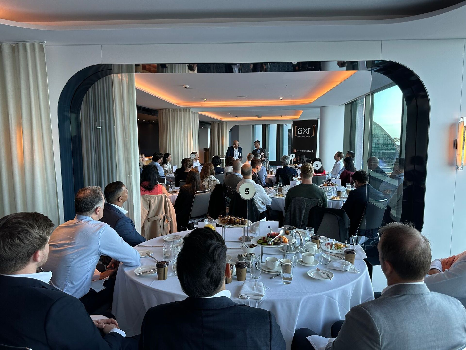 People seated at round tables in a restaurant, a speaker at the front.