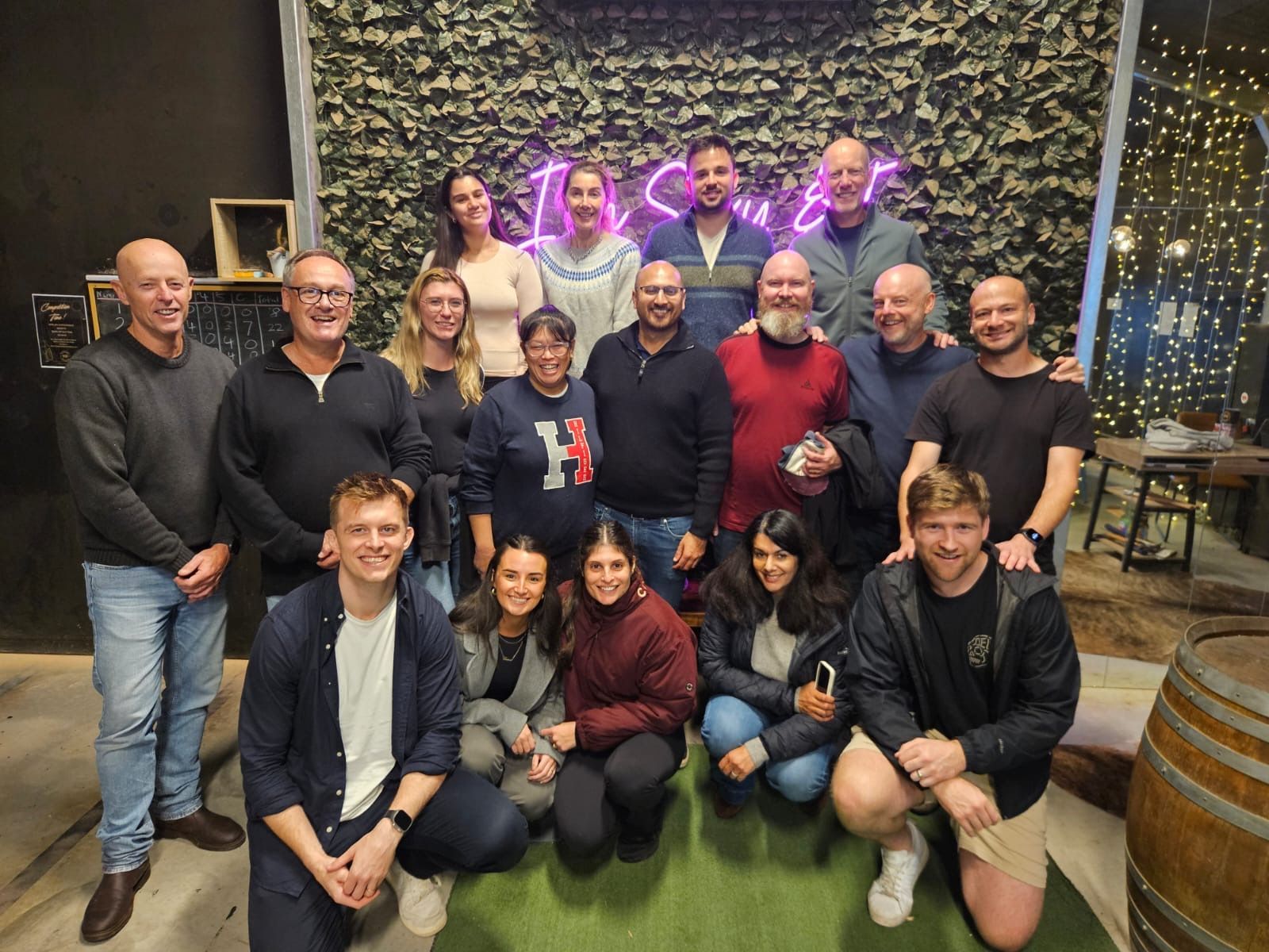 Group of people posing for a photo in front of a neon sign and a wall of greenery.