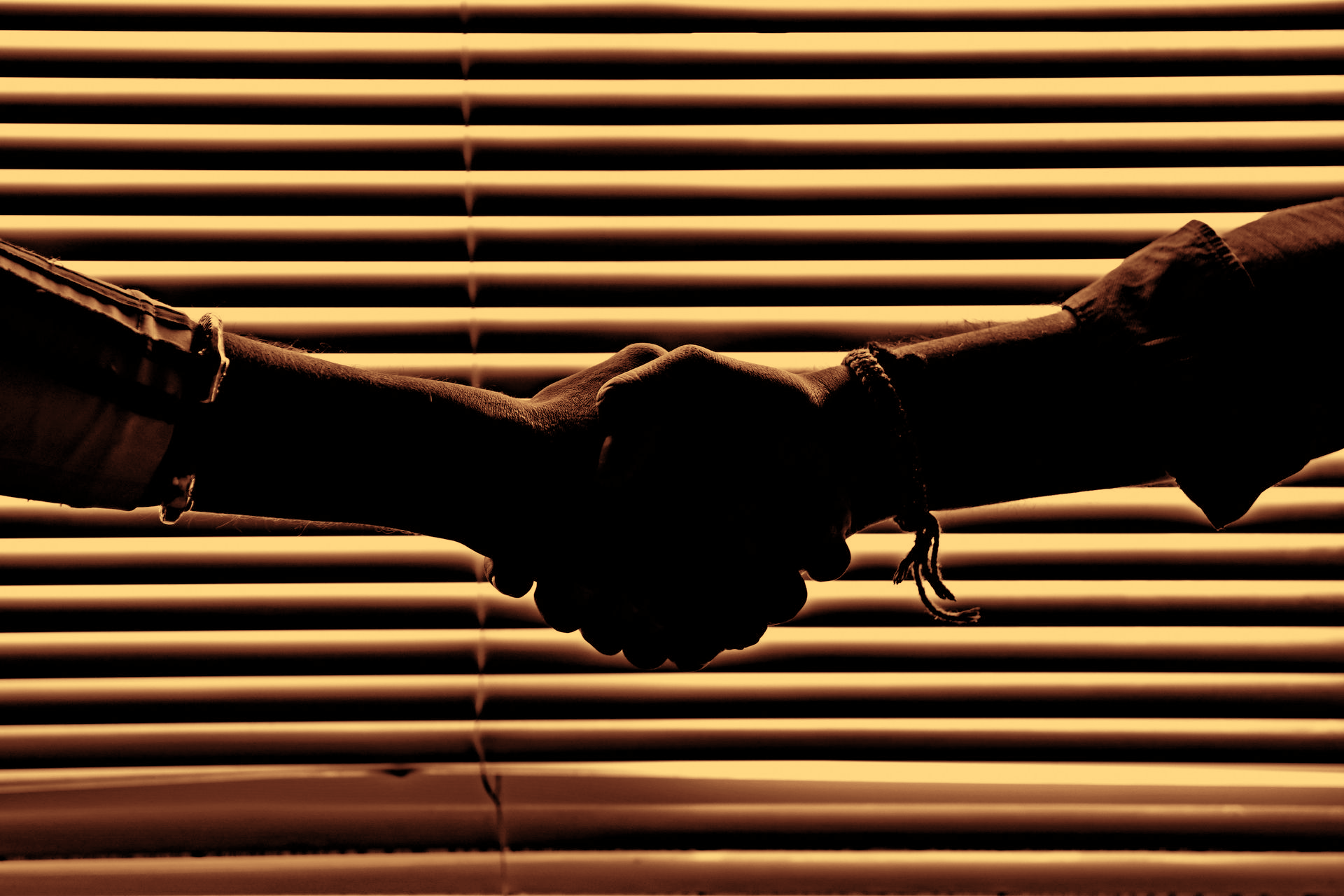 Two silhouetted arms shaking hands in front of a window with horizontal blinds.