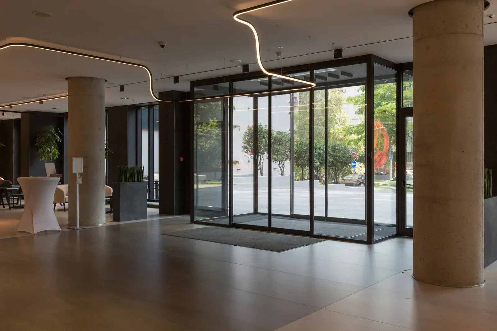 Hotel lobby with large glass doors, gray columns, and a reflective floor. Natural light streams in.