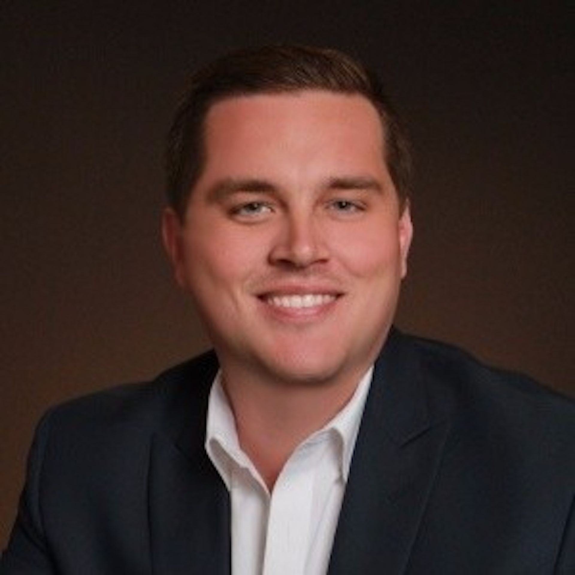 Man in a dark suit smiles at the camera, against a dark gradient background.