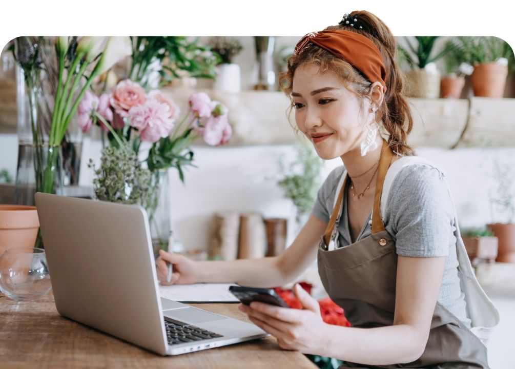 Florist working on a laptop, holding a phone, in a flower shop with floral arrangements in the background.
