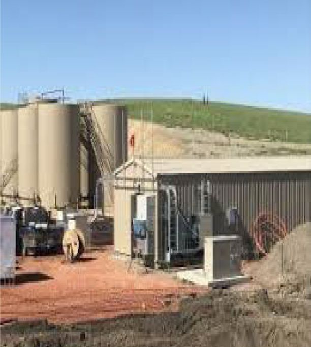 A large building with a lot of silos in the background is sitting on top of a dirt field.