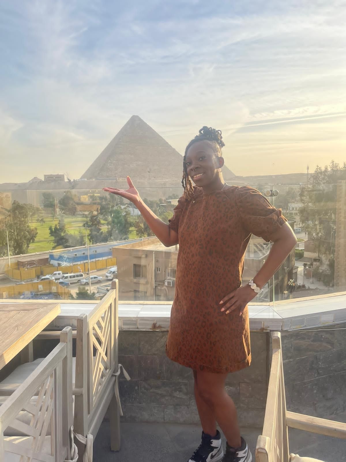 A woman in a brown dress is standing on a balcony with a pyramid in the background.