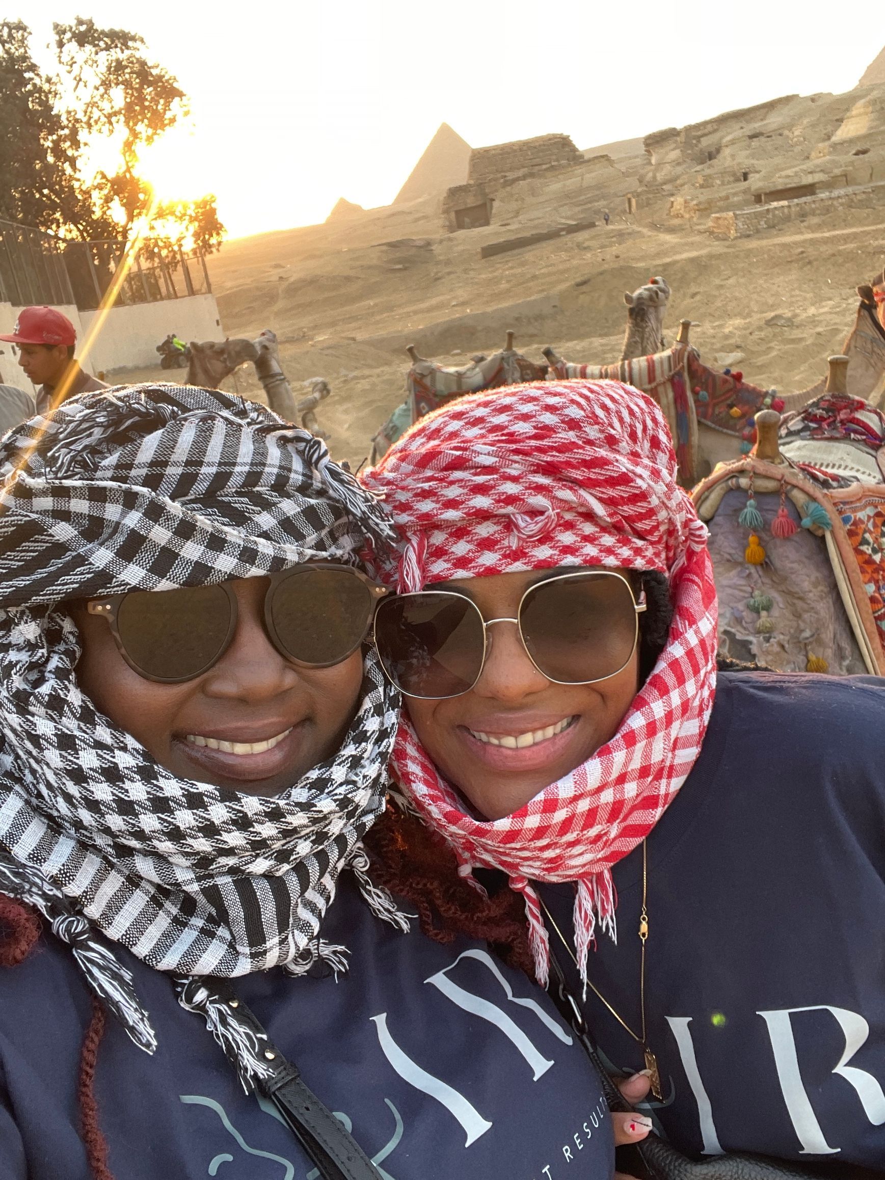 Two women wearing scarves and sunglasses are posing for a picture in front of the pyramids.