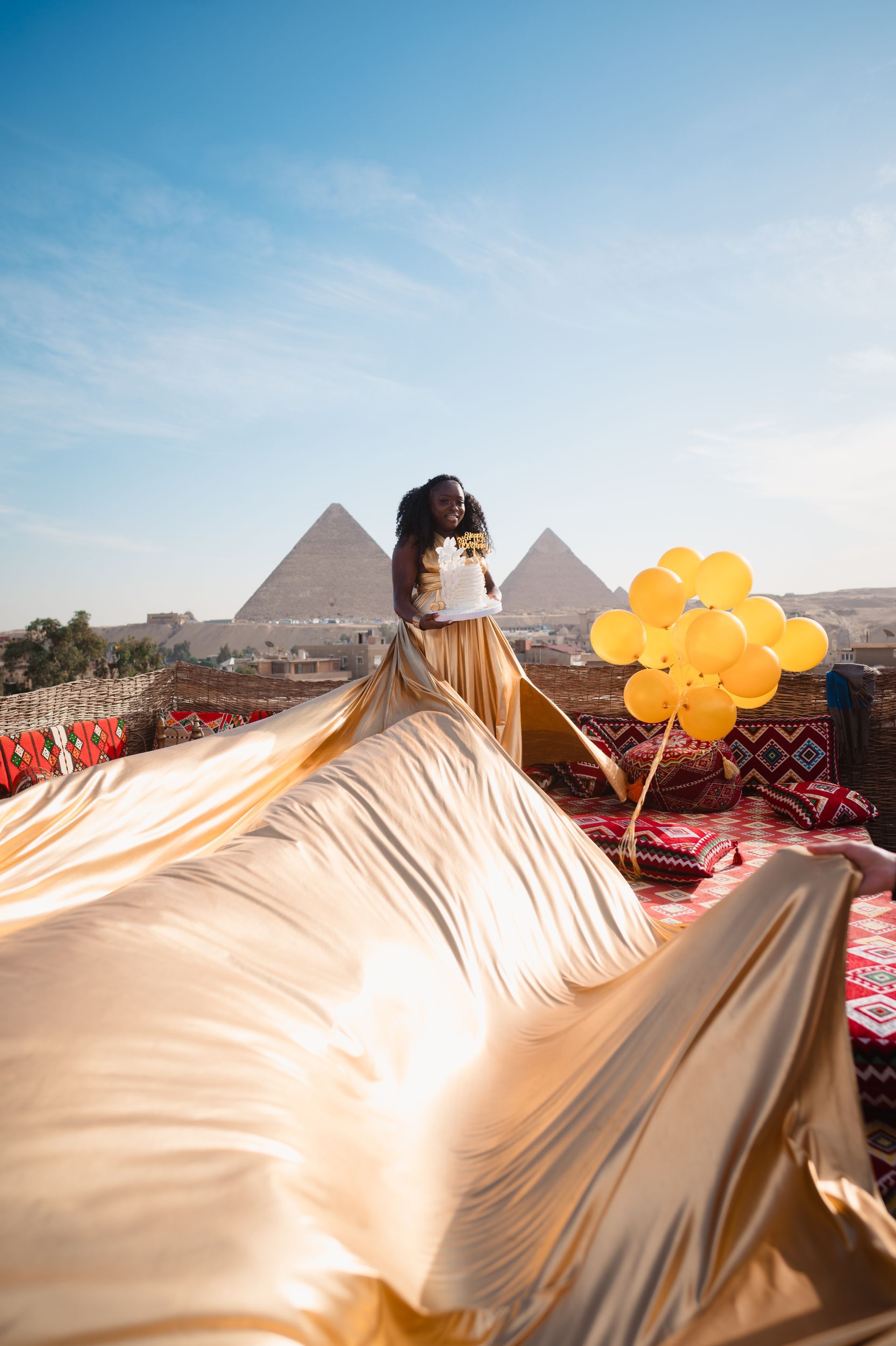 A woman in a long gold dress is standing in front of the pyramids holding balloons.