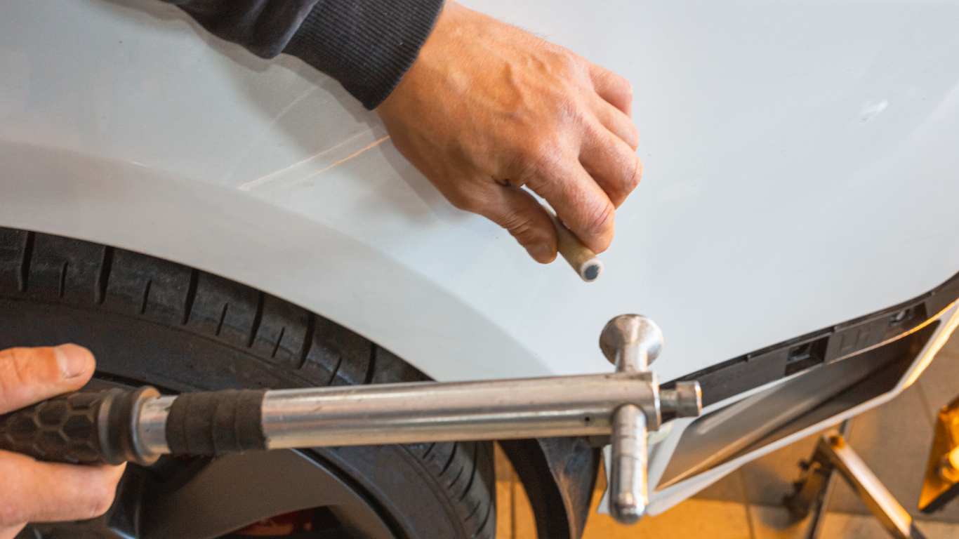 Person using a hammer and tool to repair a dent on a white car fender.