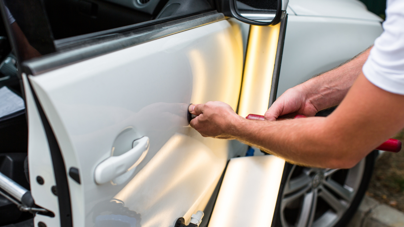A person using tools to repair a dent on a white car door, outdoors.