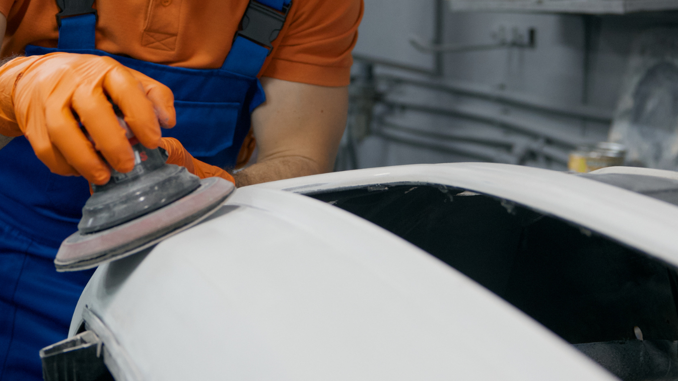 Person in orange gloves and blue overalls sanding a car part, white in color.