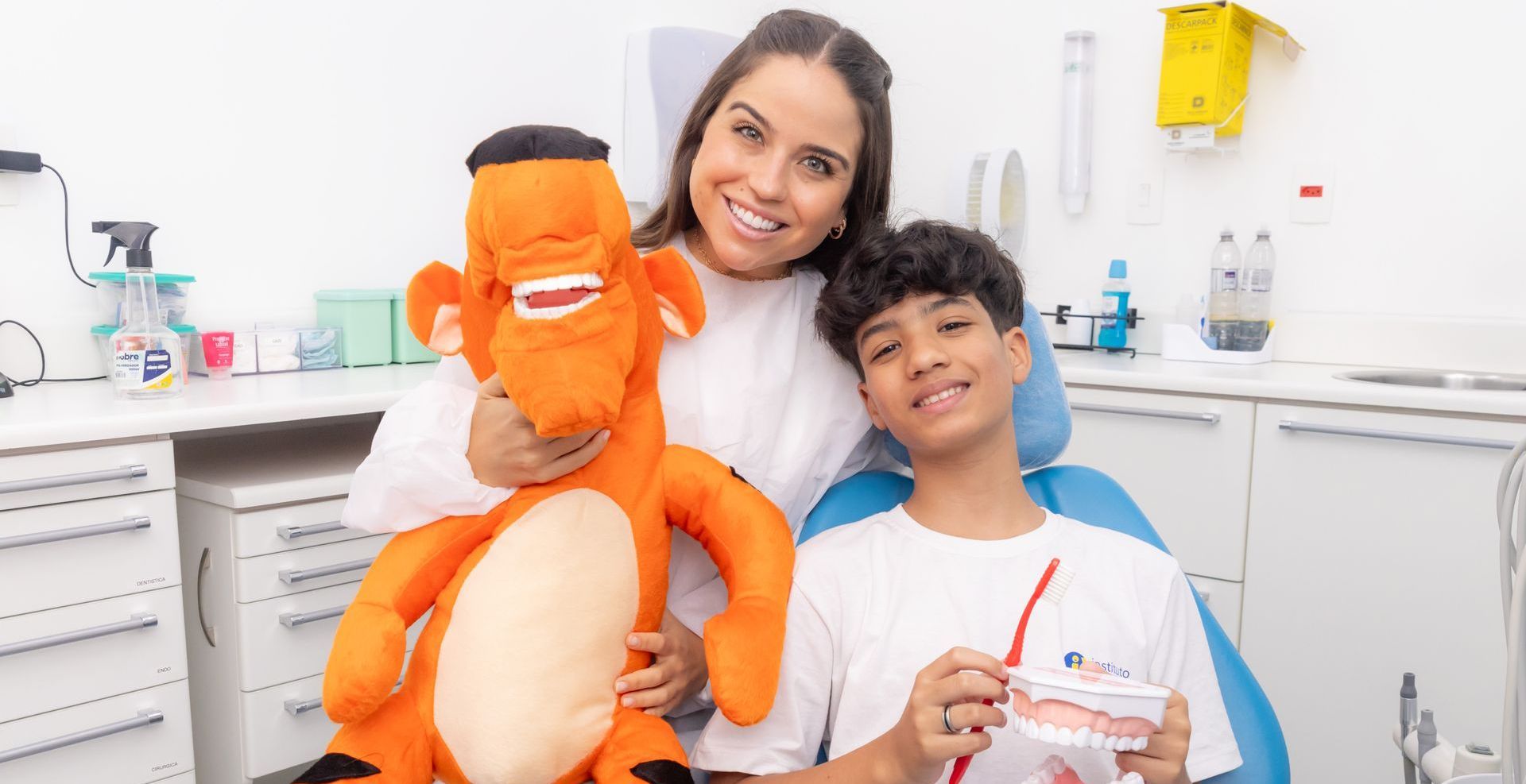 Uma mulher está segurando um bicho de pelúcia e um menino está segurando uma escova de dentes em um consultório odontológico.