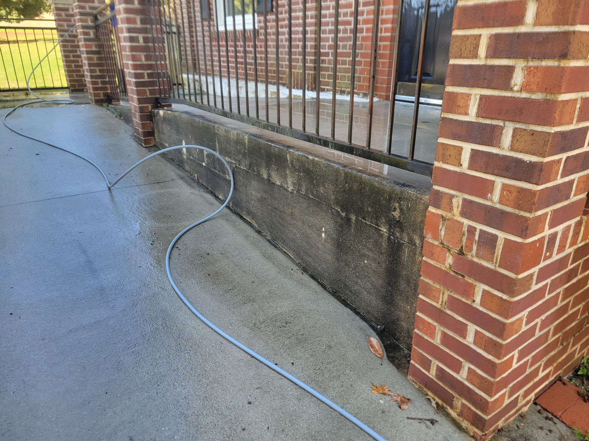 A concrete ramp being power washed with water hose next to brick columns and a wrought iron fence.