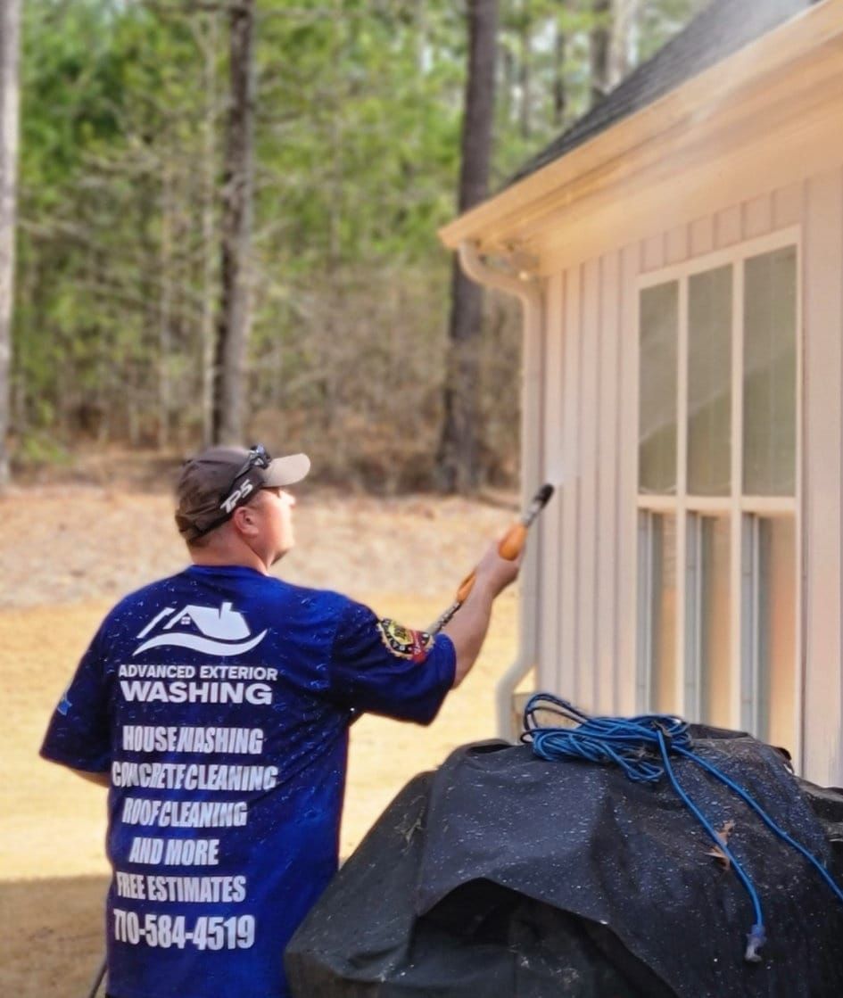 Person in yellow rain gear pressure washing a gray house with two windows.