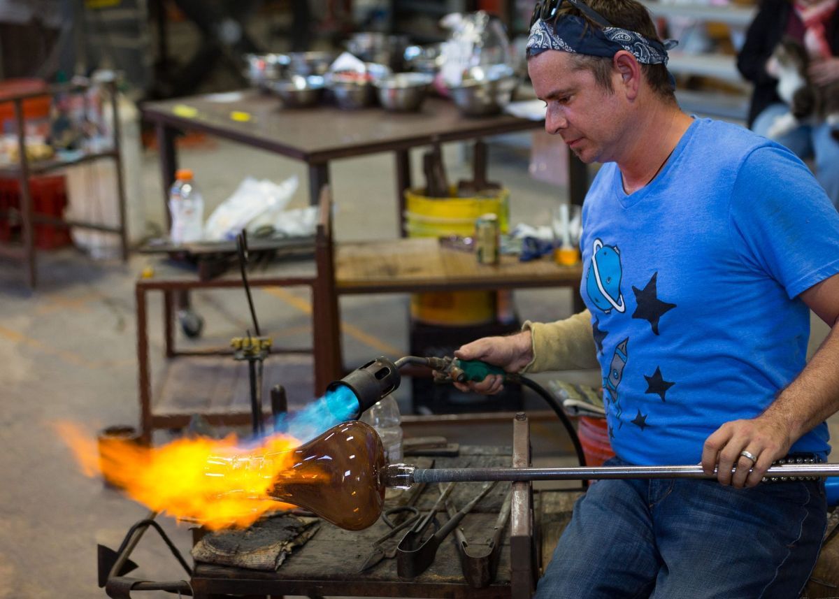 A man in a blue shirt is blowing glass with a torch.