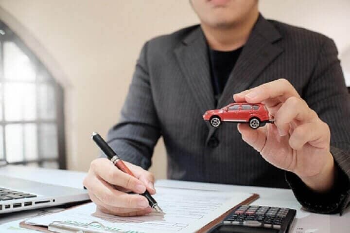 Un hombre está sentado en un escritorio sosteniendo un auto de juguete y escribiendo en un portapapeles.