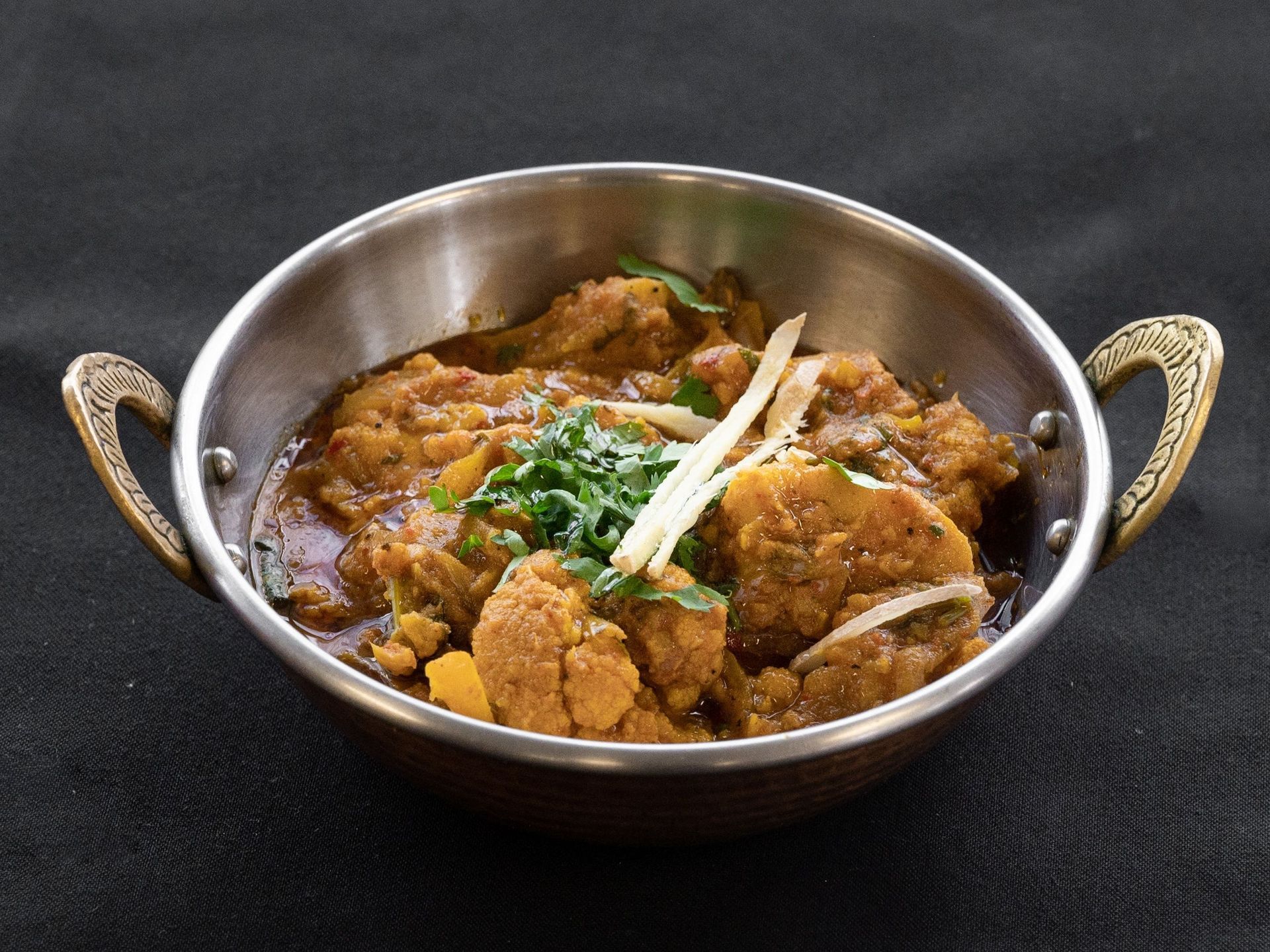 Close-up of cauliflower curry in a decorative metal bowl, garnished with ginger and cilantro.