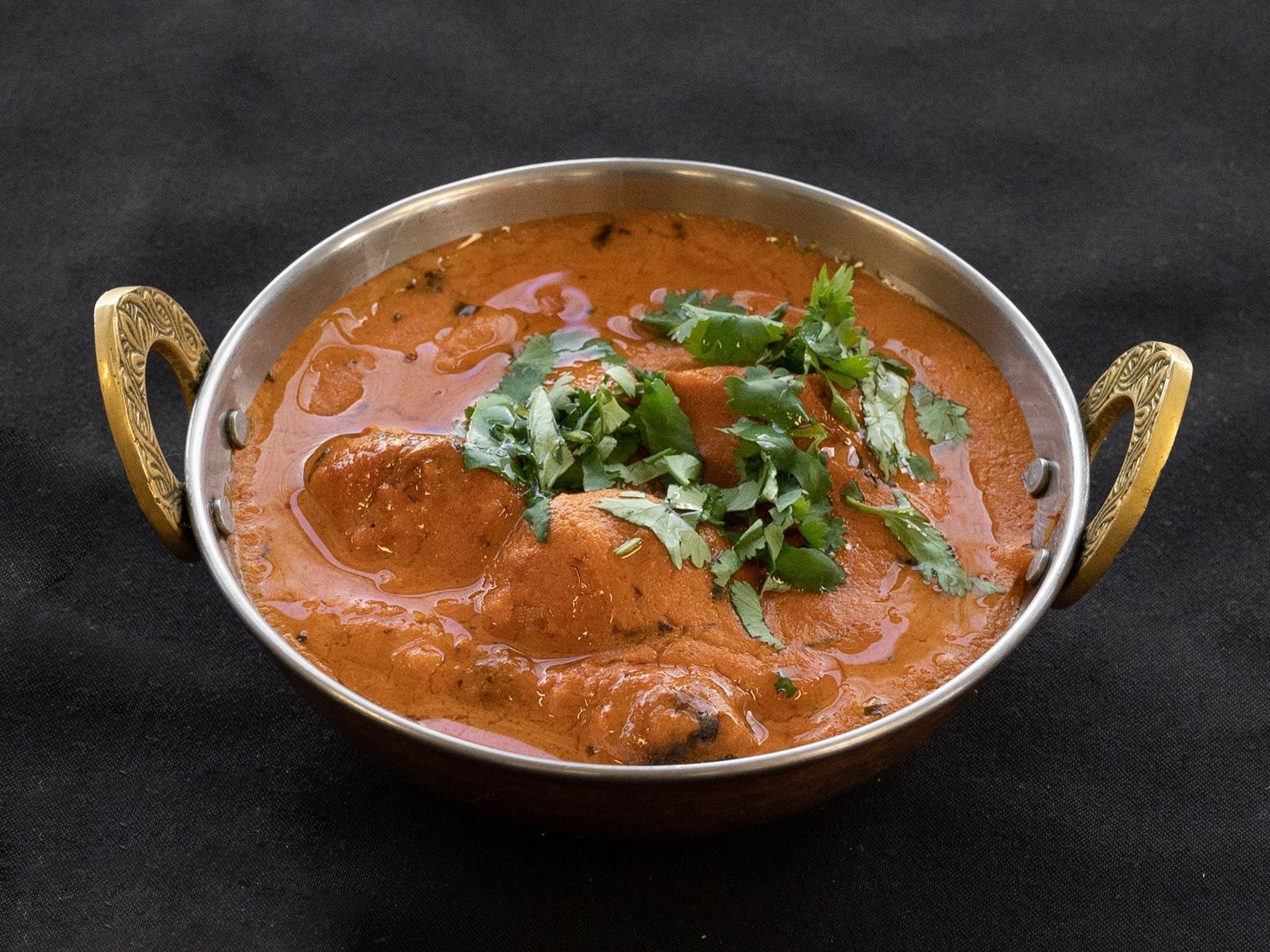 Chicken tikka masala in a copper bowl, garnished with cilantro on a black surface.