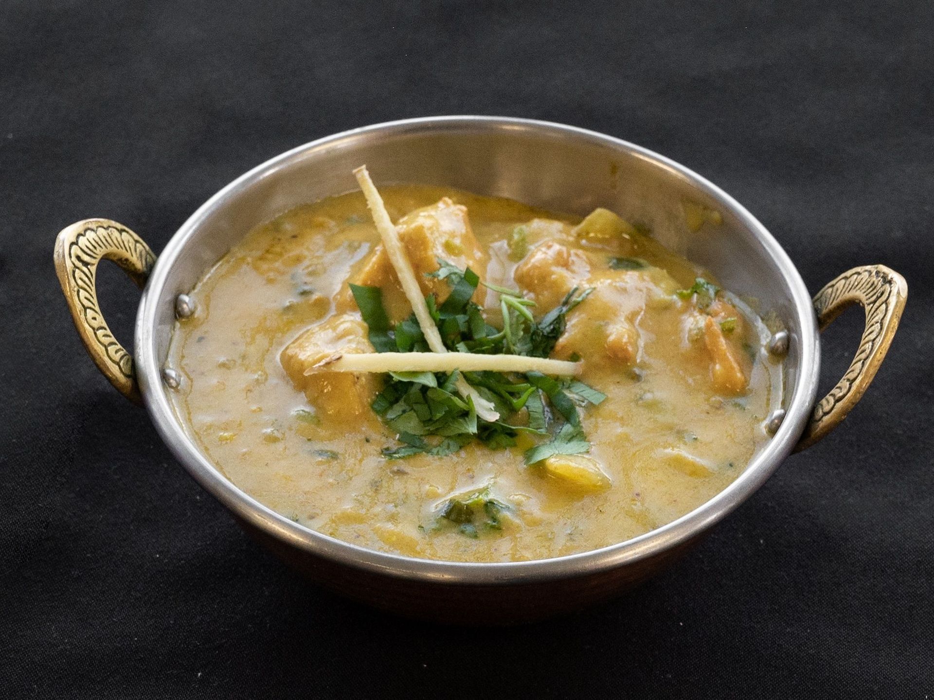Chicken curry in a decorative metal bowl, garnished with cilantro and ginger on a black surface.