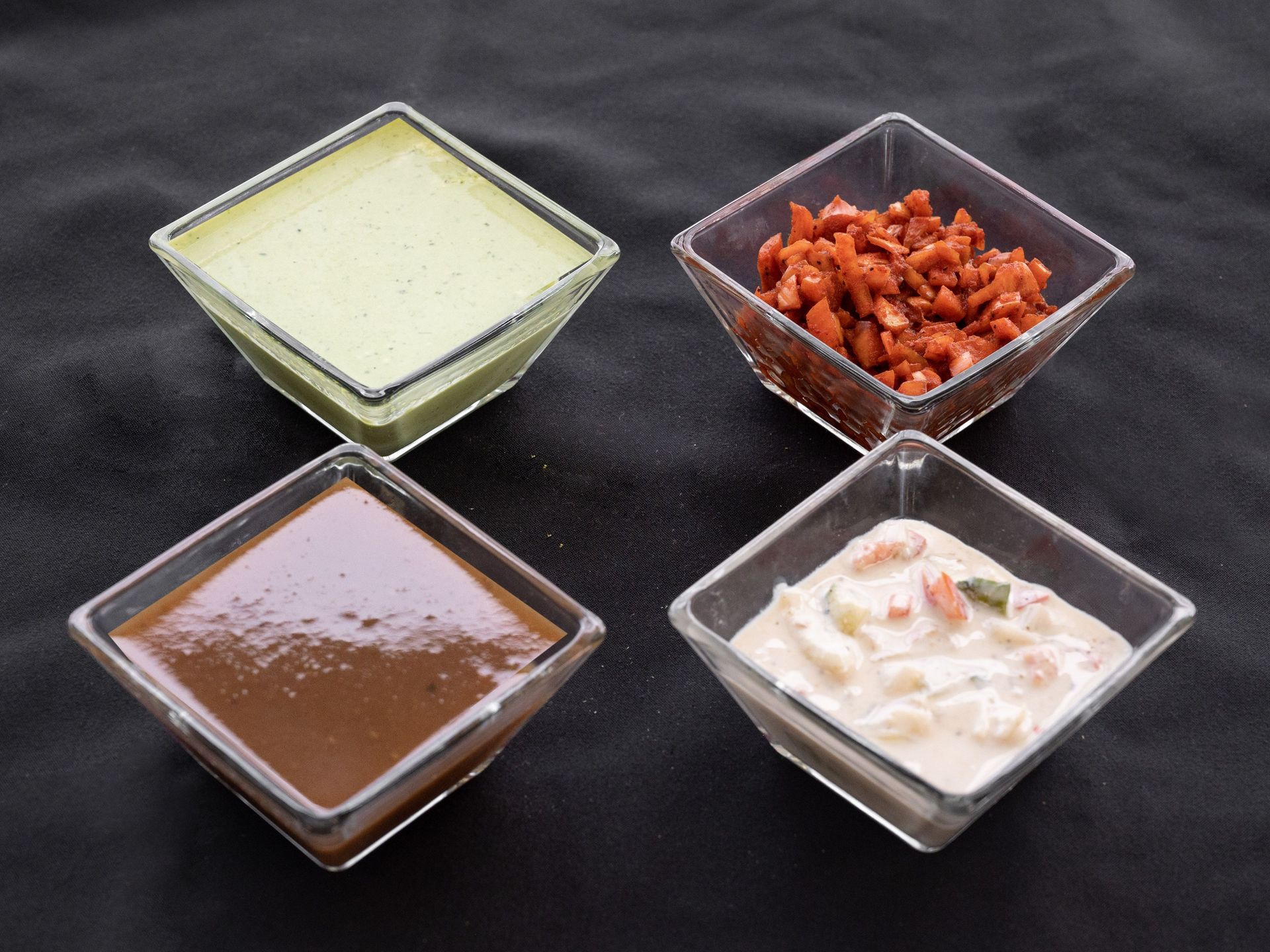 Four small square glass bowls filled with different sauces on a dark surface.