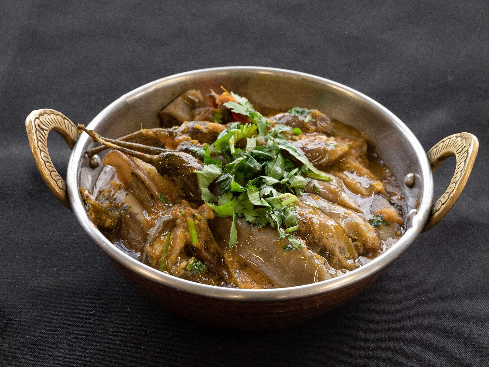 Eggplant curry in a copper bowl, garnished with cilantro, on a dark background.