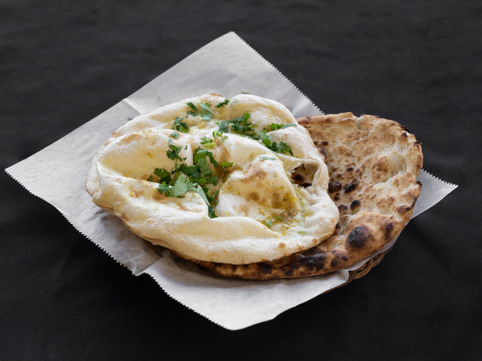 Naan bread on parchment paper, one topped with herbs, against a black background.