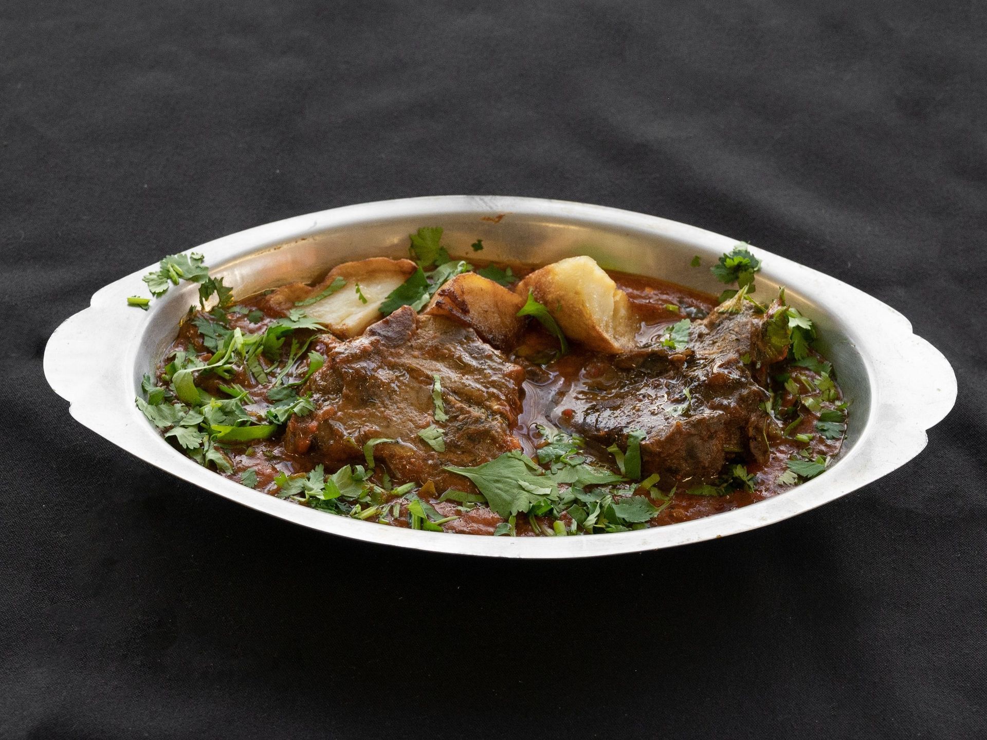 Meat curry with potatoes and cilantro in a silver dish.