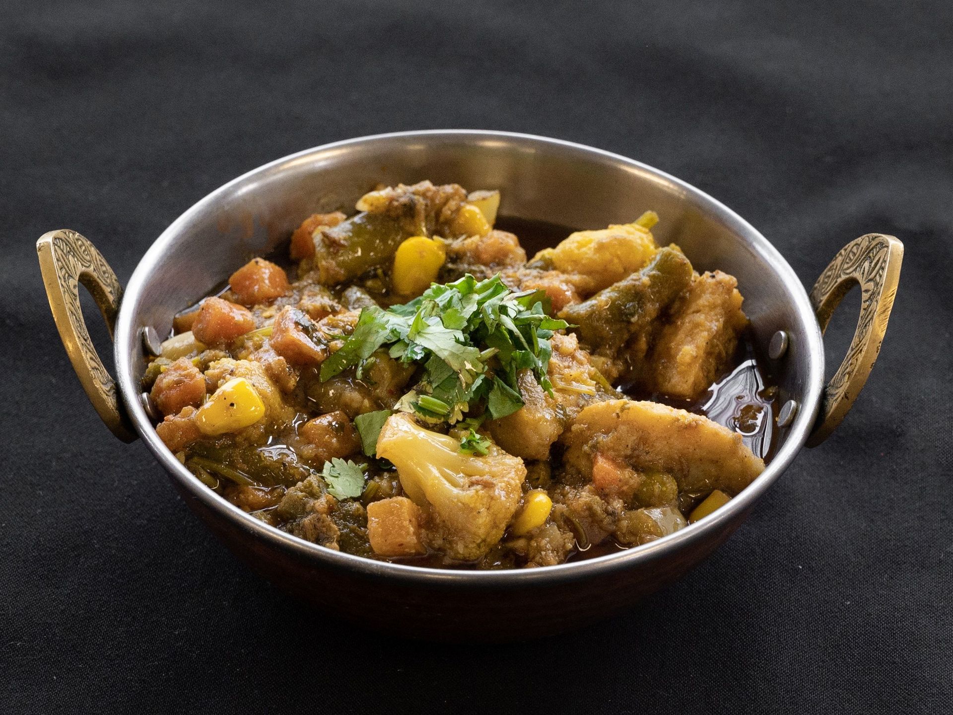 Vegetable curry in a copper bowl, garnished with cilantro, on a black surface.