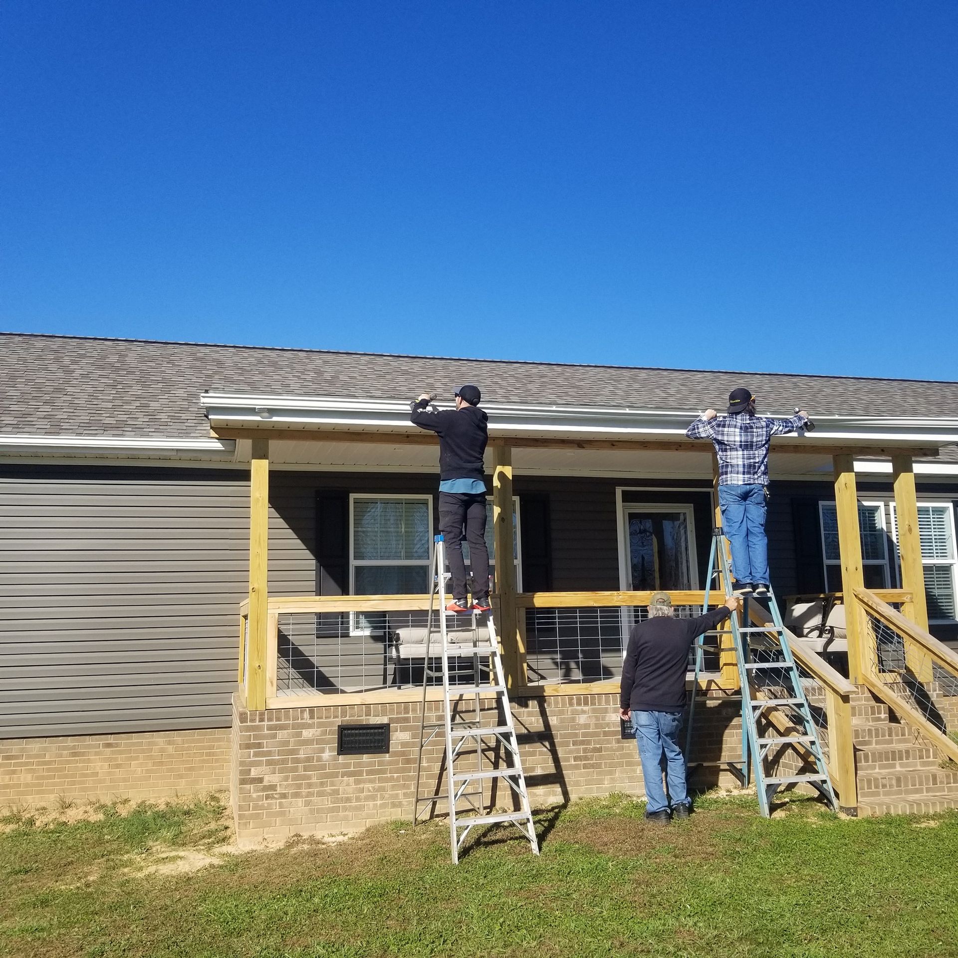 Three people on ladders working on a house porch, under a bright blue sky.